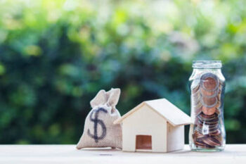 US dollar in a money bag, small residential, house model on table against green nature background.